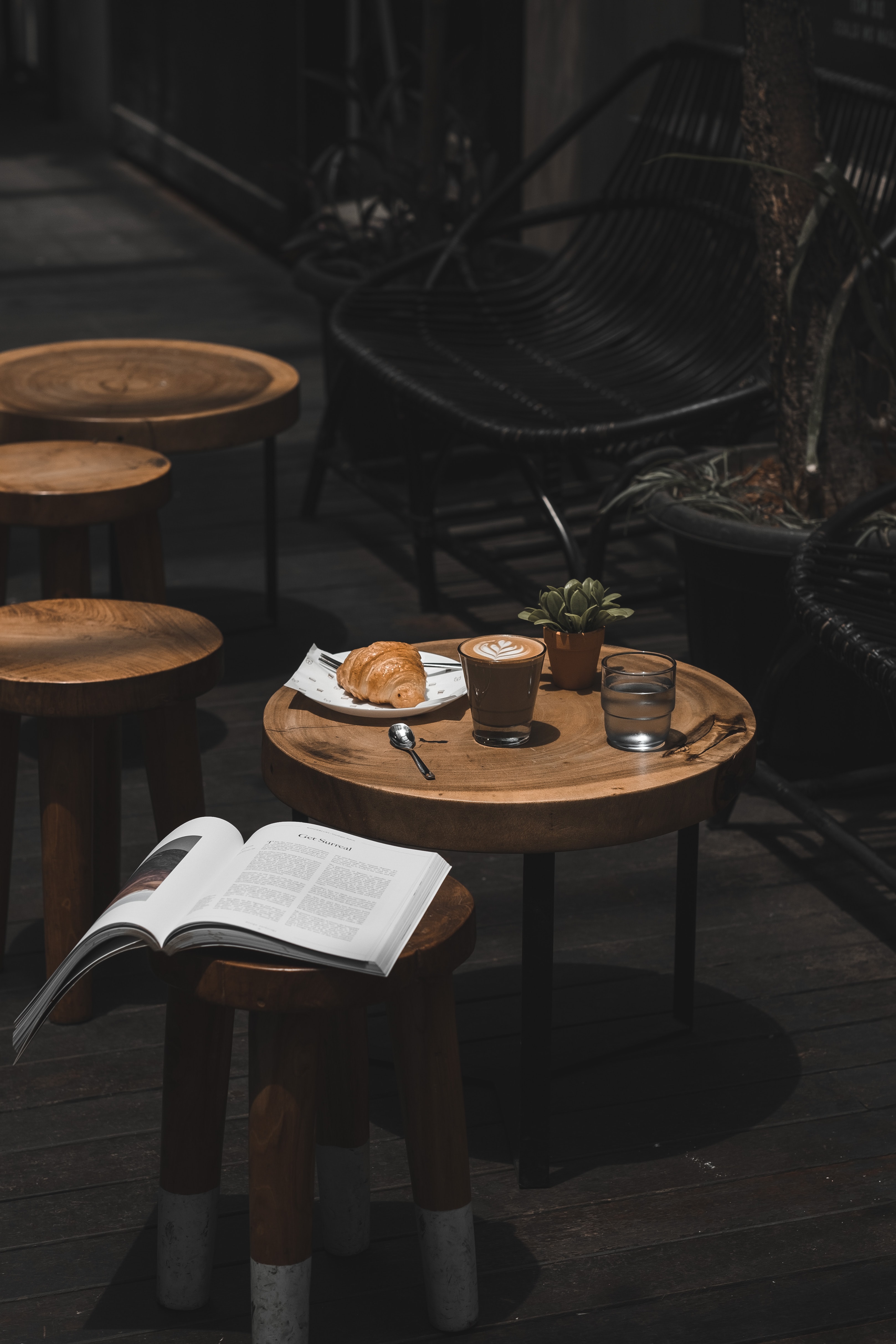 coffee shop chair and tables with a textbook on one table, and a crossaint, coffee, and plants on another.